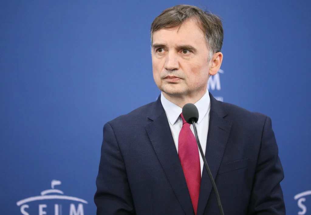 Mężczyzna w garniturze i czerwonym krawacie stoi przy mikrofonie na tle niebieskiej ściany z napisem SEJM.