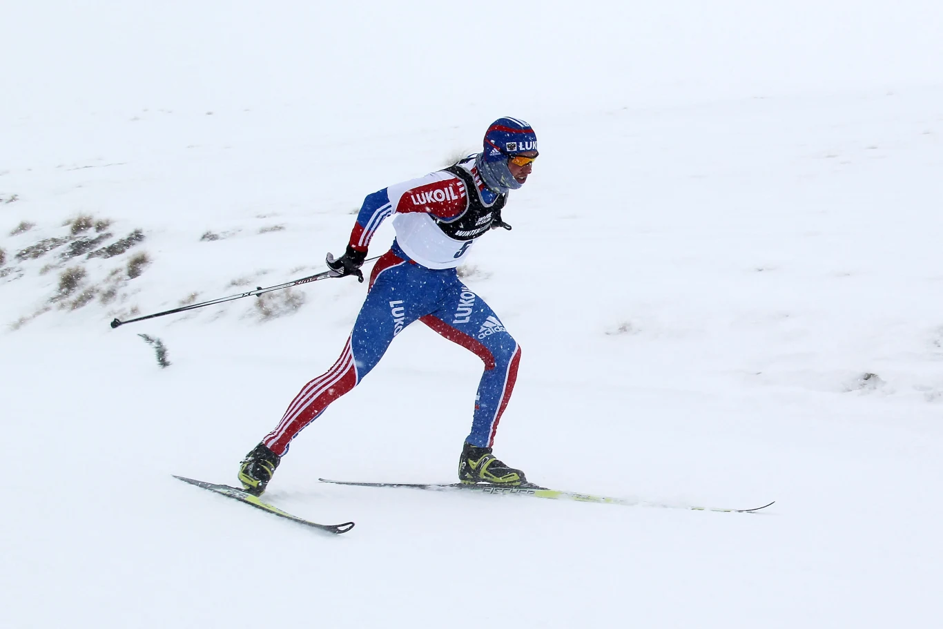 Narciarz biegowy w stroju sportowym podczas wyścigu na zaśnieżonym terenie, dynamiczna pozycja z kijkami, ślady nart widoczne na śniegu, tło zamglone i zimowe.