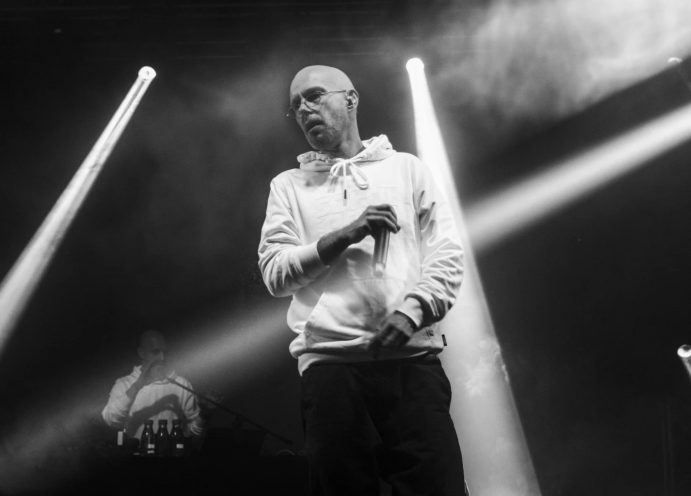 Pono miał 49 lat Mężczyzna w białej bluzie i okularach stoi na scenie, wokół niego widać światła sceniczne i dym, co wskazuje na występ muzyczny. W tle znajduje się druga postać, najprawdopodobniej DJ lub realizator dźwięku.