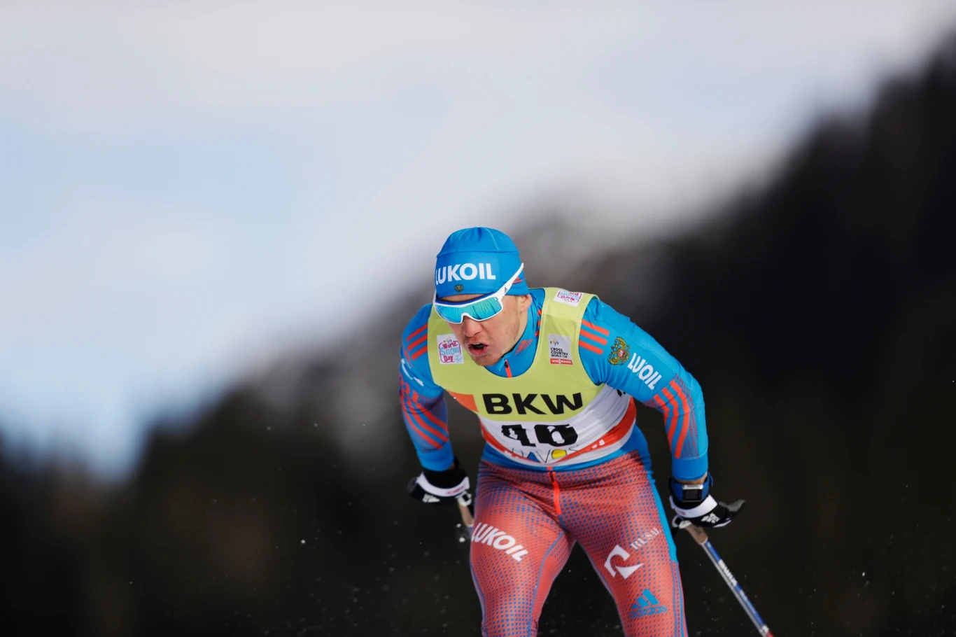 Zawodnik w stroju narciarskim i czapce startuje w biegu narciarskim, trzymając kijki, na piersi widoczny numer startowy oraz logo sponsorów. W tle rozmyte drzewa i góry.