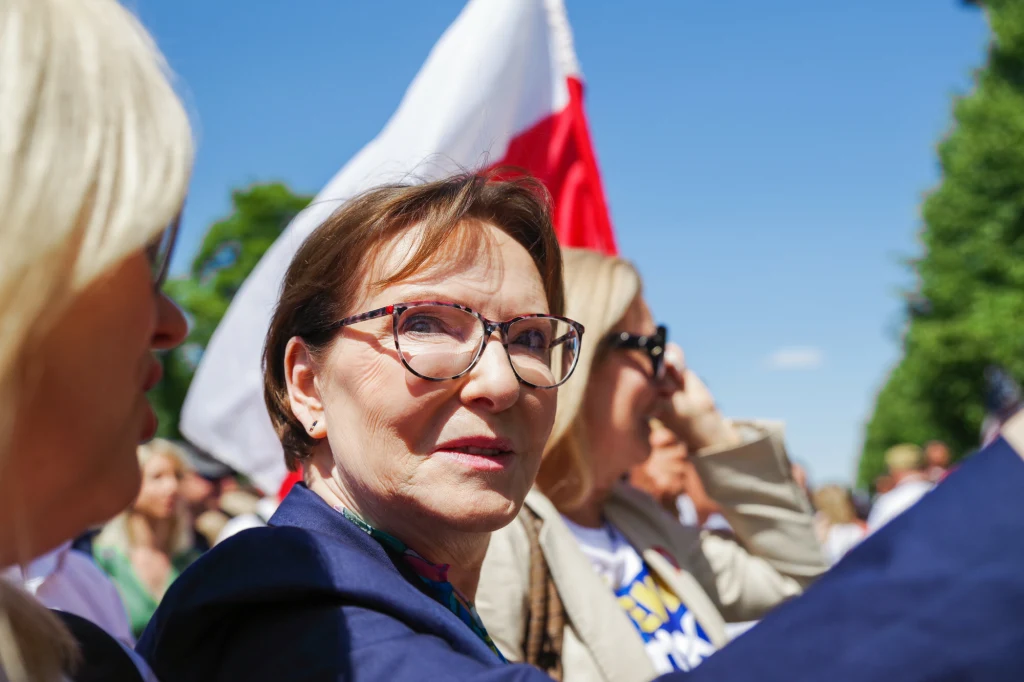 Europosłanka Koalicji Obywatelskiej i była premier Ewa Kopacz Kobieta w okularach na pierwszym planie, uśmiechnięta, otoczona innymi osobami, w tle polska flaga oraz jasnoniebieskie niebo, atmosfera wydarzenia plenerowego.
