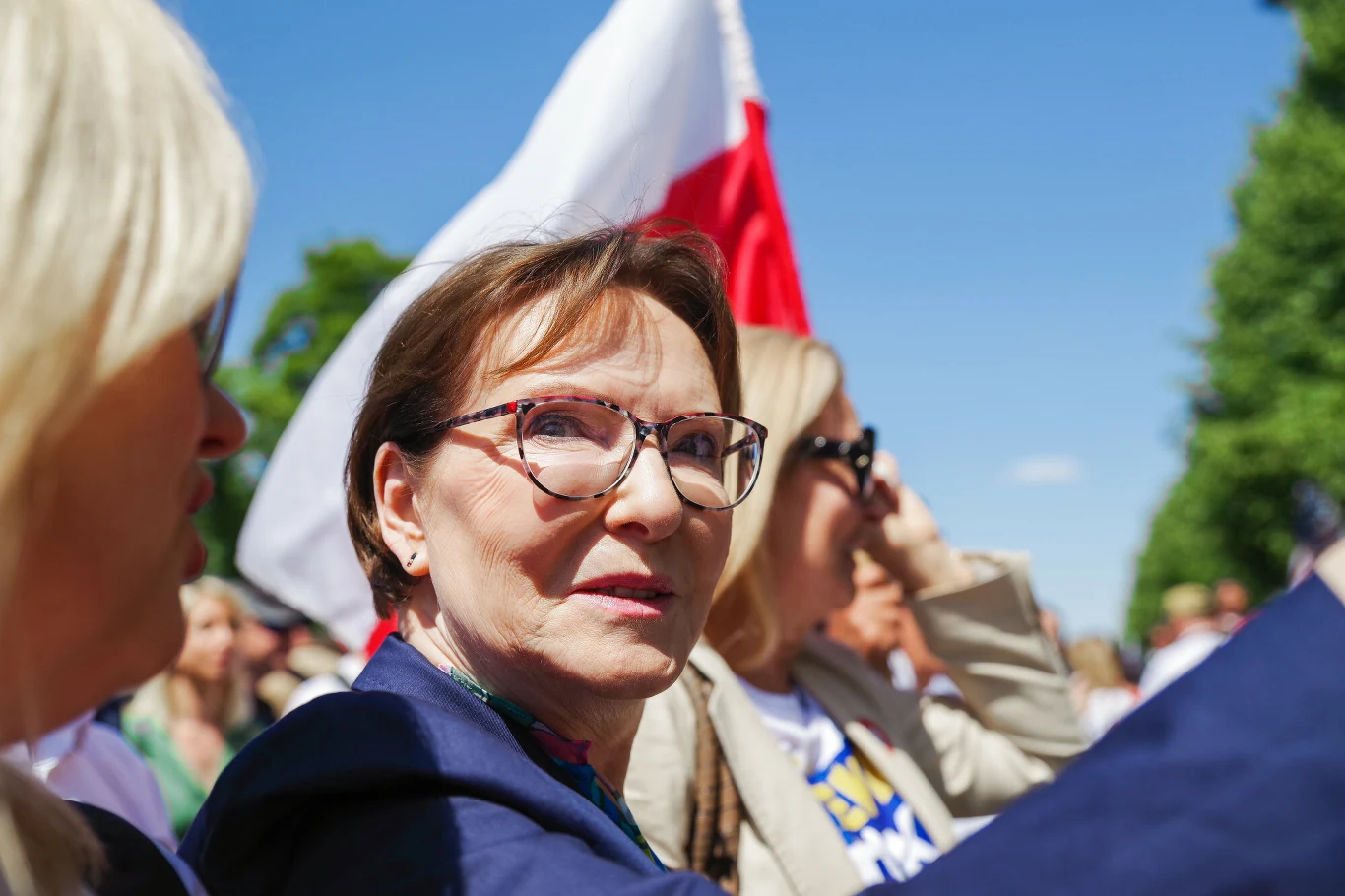 Europosłanka Koalicji Obywatelskiej i była premier Ewa Kopacz Kobieta w okularach na pierwszym planie, uśmiechnięta, otoczona innymi osobami, w tle polska flaga oraz jasnoniebieskie niebo, atmosfera wydarzenia plenerowego.