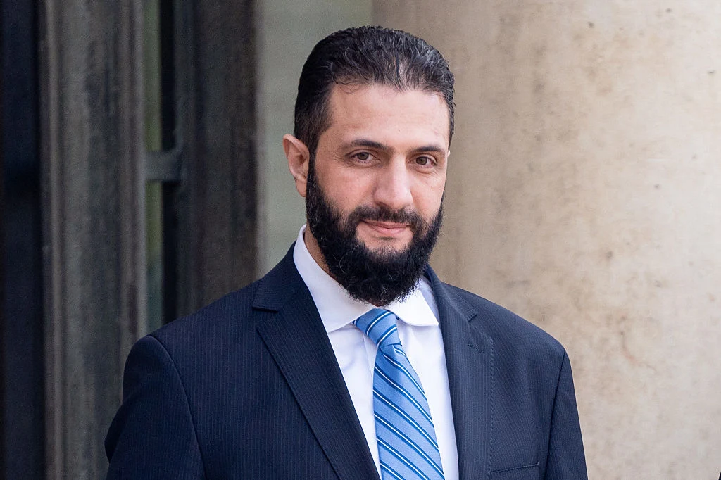 Prezydent Syrii Ahmed al-Sharaa odwiedzi w poniedziałek Biały Dom Mężczyzna w garniturze i niebieskim krawacie stoi na tle jasnej ściany i ciemnych drzwi, patrząc bezpośrednio w stronę aparatu.