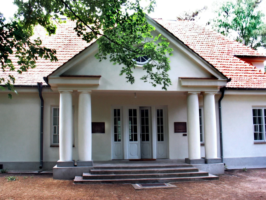 Klasyczny dworek z białą elewacją, kolumnowym gankiem oraz czerwonym dachem, otoczony przez drzewa i niewielki ogród. Stromy dach, symetryczne okna i schody prowadzące do wejścia tworzą harmonijną całość architektoniczną.