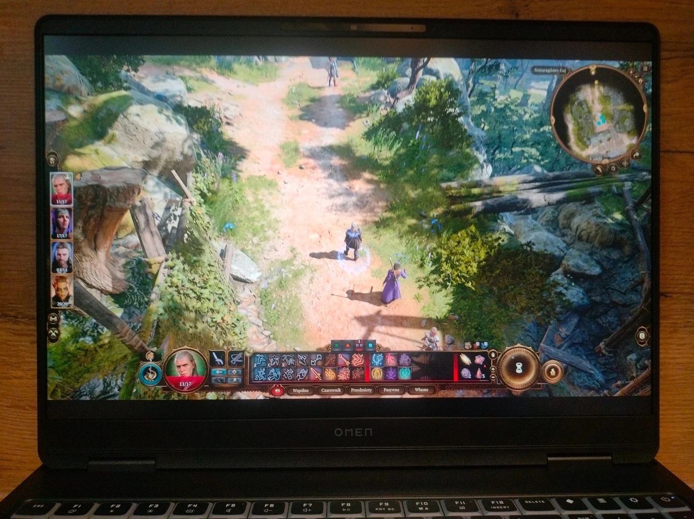 Ekran laptopa wyświetlający rozgrywkę w grę komputerową typu RPG, z widokiem izometrycznym na drużynę postaci przemierzających leśną ścieżkę, widoczny interfejs gry oraz lista bohaterów z boku ekranu.