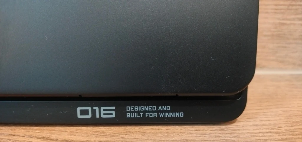 OMEN MAX 16 Fragment czarnej obudowy z widocznym napisem '016 Designed and built for winning' na tle drewnianego podłoża.
