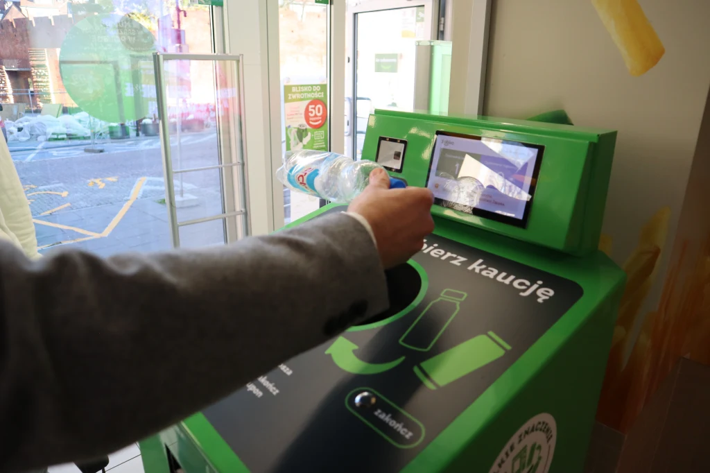 Do obsługi butelkomatów w Żabce nie potrzeba aplikacji. Maszyna sama sprawdza, czy skanowane opakowania są objęte kaucją, czy nie Osoba wkłada plastikową butelkę do zielonego automatu do zwrotu opakowań ustawionego w sklepie. Na urządzeniu widoczne są ilustracje instruktażowe, a obok wejście do sklepu.