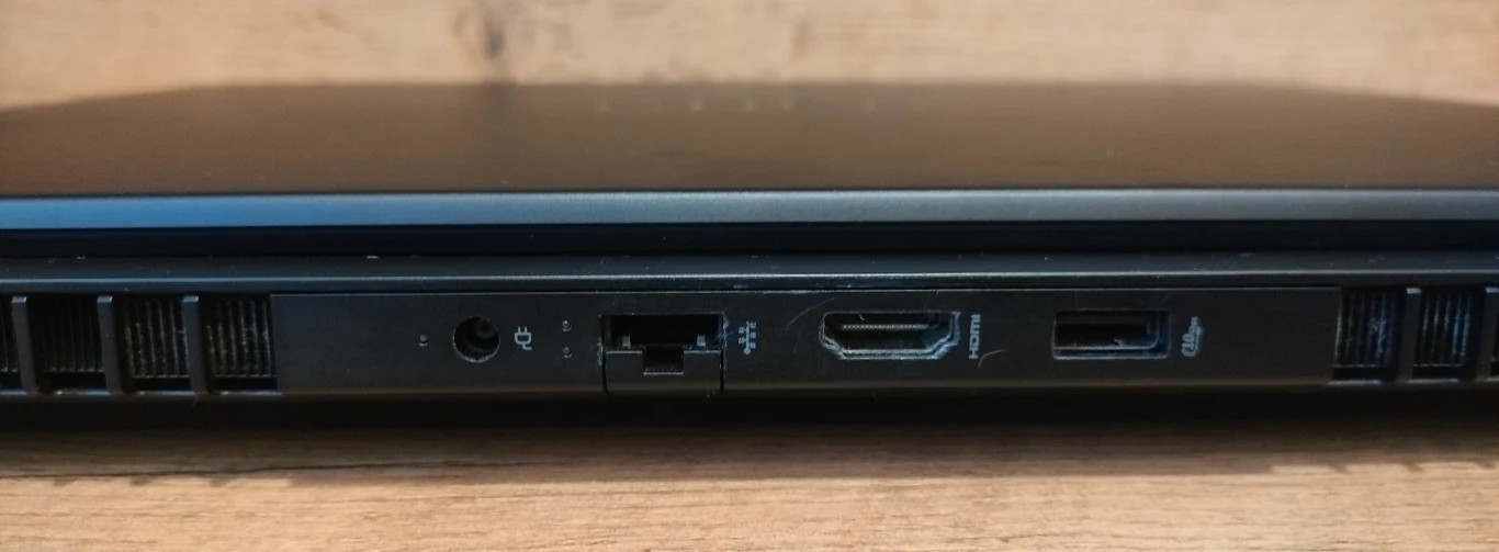 Boczna krawędź laptopa z widocznymi portami, w tym wejściem słuchawkowym, portem ethernetowym, HDMI i USB, na tle drewnianego blatu.