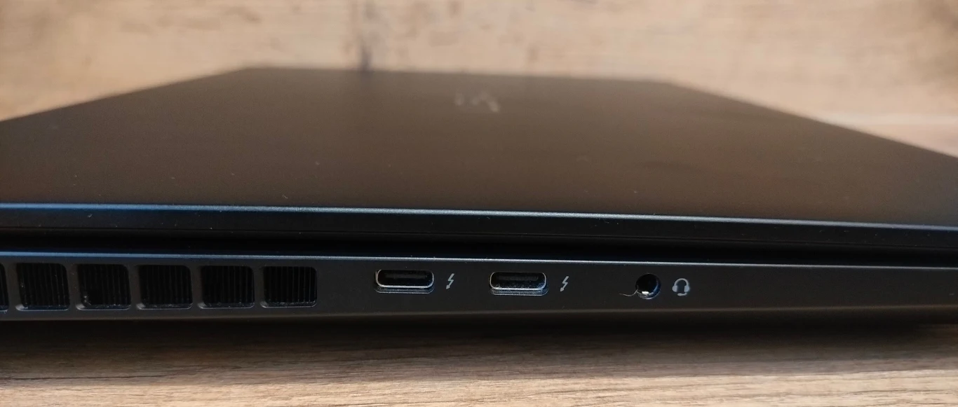 Czarny laptop ustawiony na drewnianej powierzchni, widoczny bok urządzenia z dwoma portami USB typu C ze znakiem błyskawicy, portem audio oraz otworami wentylacyjnymi po lewej stronie.