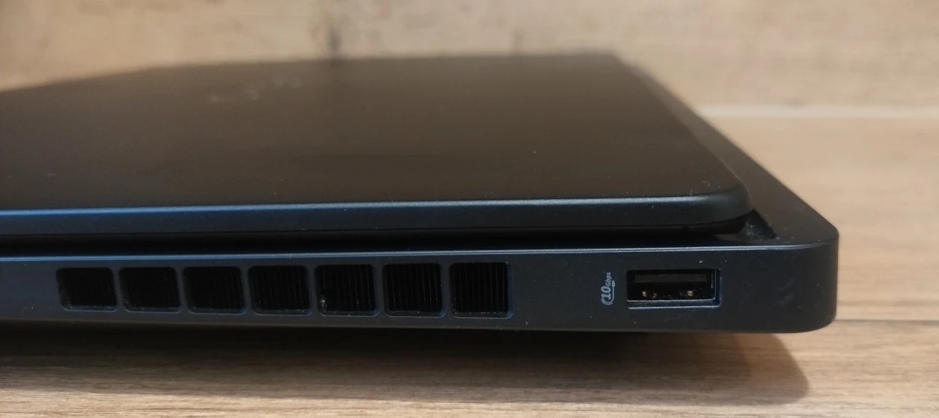 Czarna obudowa zamkniętego laptopa z widocznym wylotem wentylacyjnym oraz portem USB umieszczonym z boku, całość ustawiona na drewnianej powierzchni.