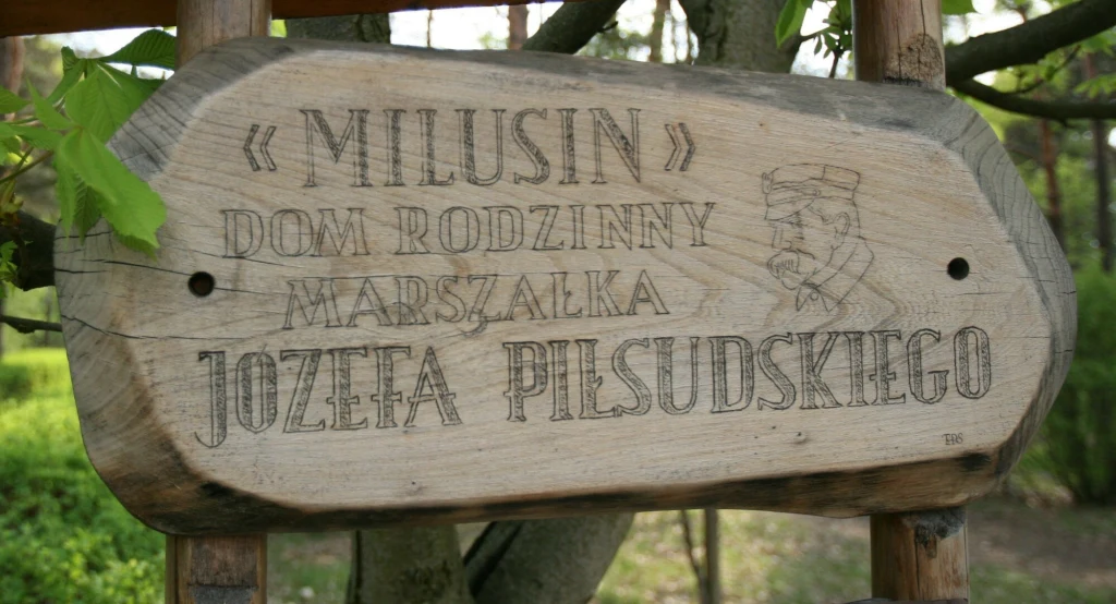Drewniana tablica z napisem „Milusin” dom rodzinny Marszałka Józefa Piłsudskiego zawieszona na tle drzewa i zieleni, z wyrytym wizerunkiem Piłsudskiego po prawej stronie.