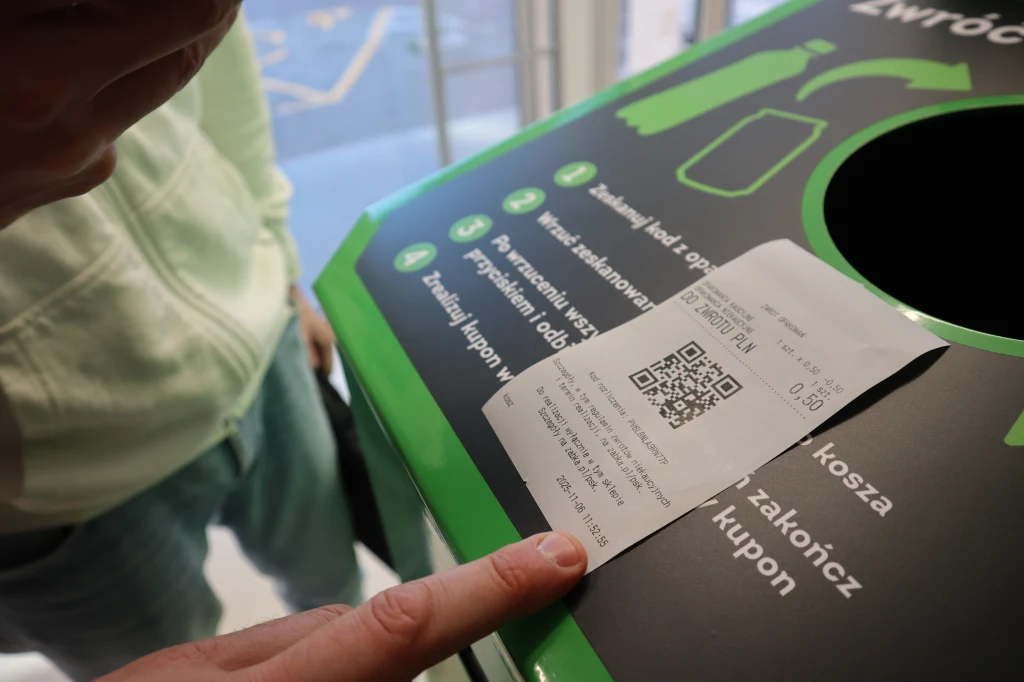 W Żabce preferowaną metodą zwrotu kaucji jest gotówka, ale można ją odebrać także w formie zniżki na zakupy Osoba w zielonej bluzie i niebieskich spodniach korzysta z automatu do zwrotu opakowań, kładzie wydrukowany kupon z kodem QR na maszynie, na której znajdują się instrukcje zwrotu oraz grafiki przedstawiające plastikowe butelki.