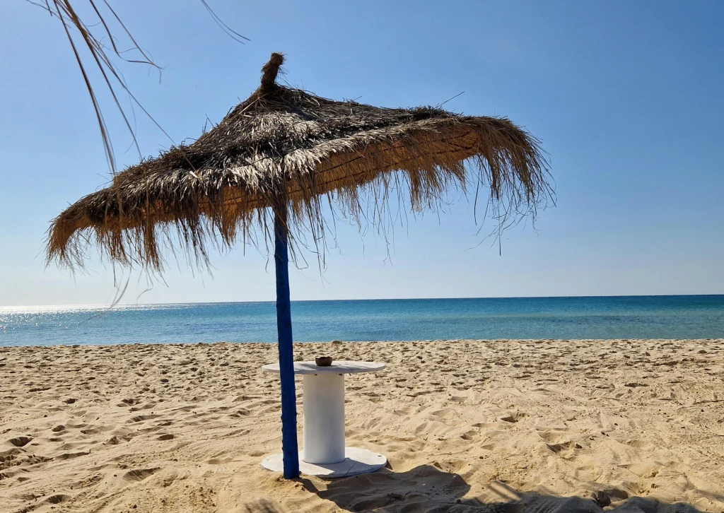 Słomiany parasol plażowy rzucający cień na piasek, pod nim niewielki okrągły stolik stojący tuż przy brzegu morza. W tle spokojna woda oraz czyste, błękitne niebo.