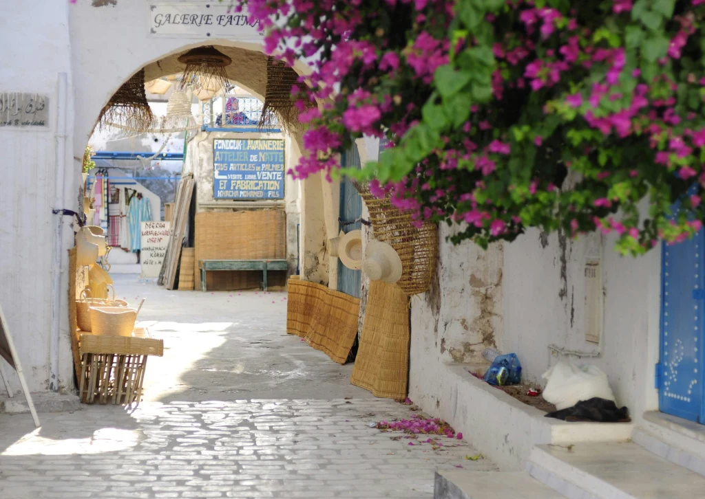 Biała, wąska uliczka z łukiem i sklepikami, udekorowana kwitnącymi różowymi bugenwillami. Po lewej stoją wiklinowe kosze i kapelusze, a po prawej stronie znajduje się niebieskie drzwi i pusta ławka.