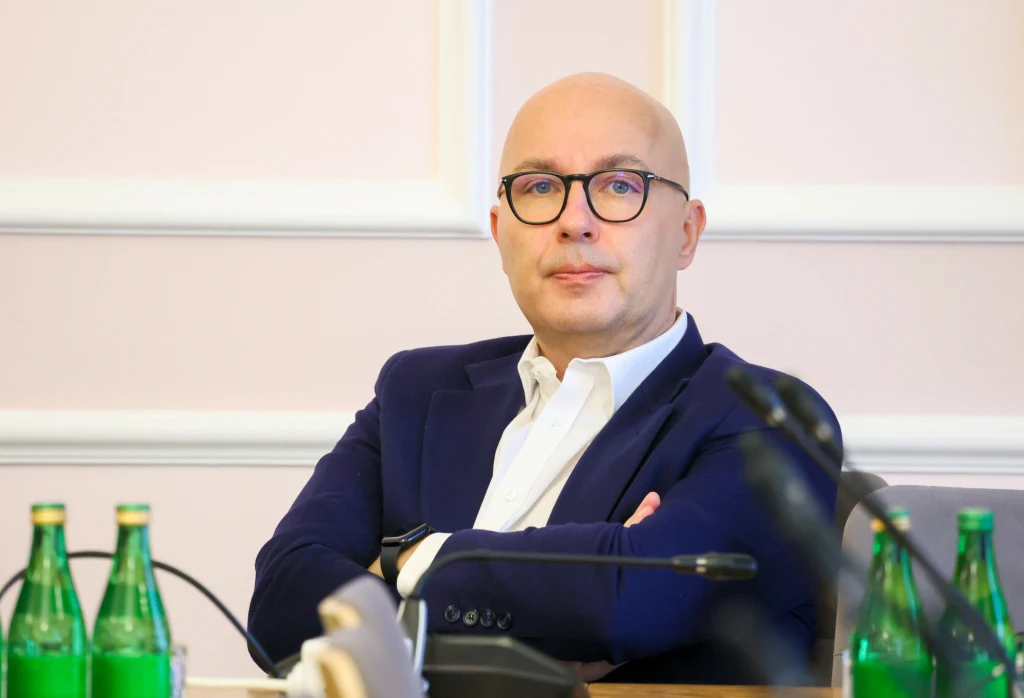 Dymisja Roberta Kropiwnickiego budzi emocje na sejmowych korytarzach Mężczyzna w okularach i garniturze siedzi przy stole konferencyjnym, na blacie kilka butelek wody mineralnej, w tle jasna ściana z białymi listwami.