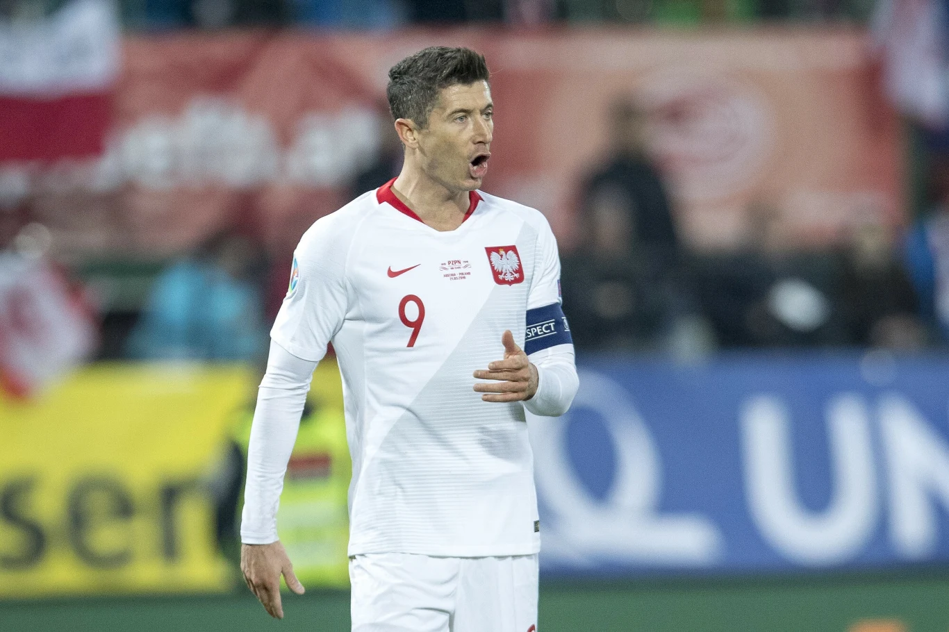 Robert Lewandowski Piłkarz w białym stroju reprezentacji Polski z numerem 9 i opaską kapitana, podczas meczu na stadionie, tło wypełniają kibice i kolorowe banery.