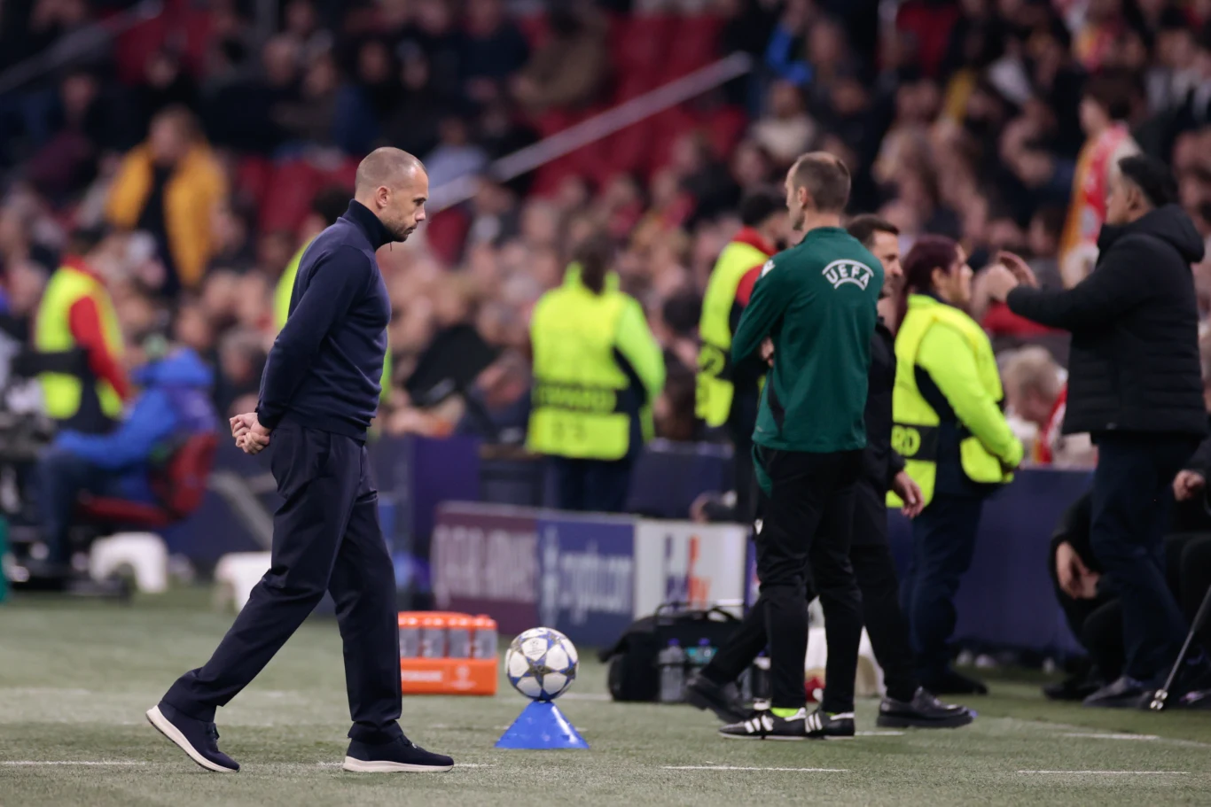 John Heitinga Trener piłkarski w ciemnym ubraniu przechadza się wzdłuż linii bocznej boiska podczas meczu. W tle widoczny sędzia UEFA oraz pozostali członkowie zespołu technicznego i ochrona. Na pierwszym planie piłka na stożku, za nią trybuny z kibicami.