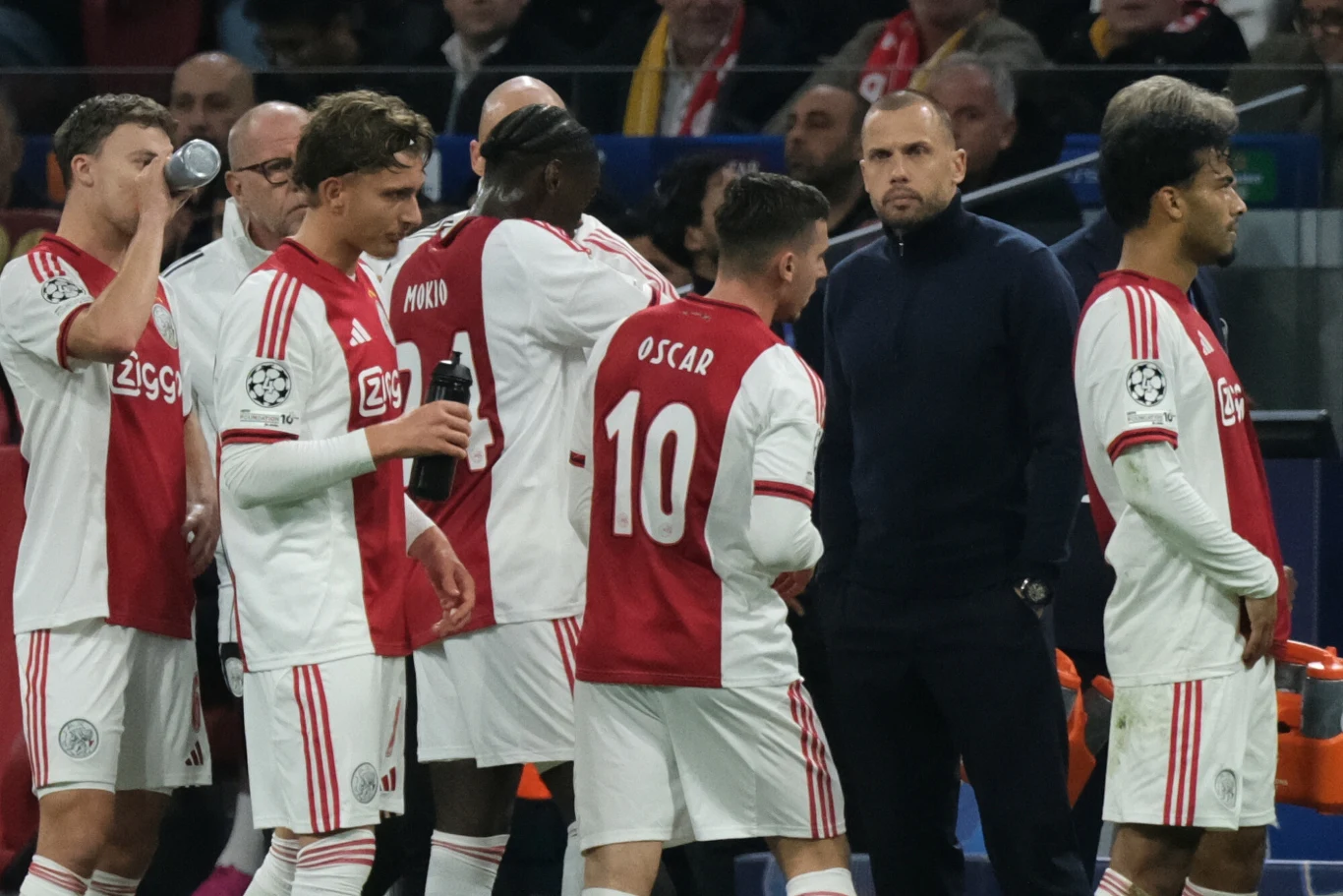Ajax Amsterdam pod wodzą Johna Heitingi zanotował fatalną serię czterech porażek z rzędu w Lidze Mistrzów Piłkarze drużyny Ajax Amsterdam ubrani w biało-czerwone stroje oraz trener stoją na boisku podczas meczu piłkarskiego, część zawodników pije wodę, atmosfera skupienia i przygotowania do gry.