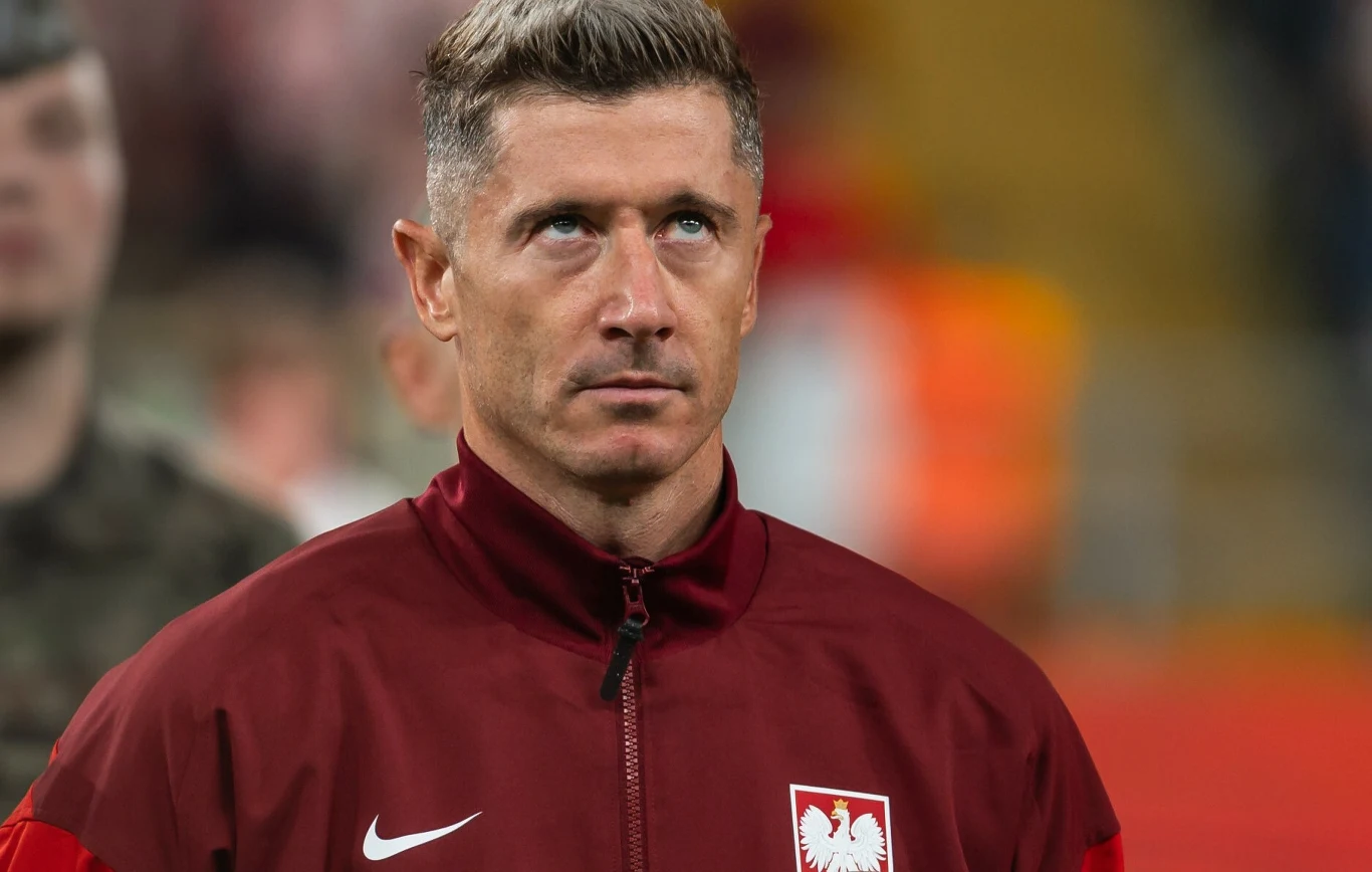 Robert Lewandowski Mężczyzna w bordowej kurtce z godłem Polski na piersi stoi na tle rozmytego stadionu, skupiony i patrzący lekko w górę.
