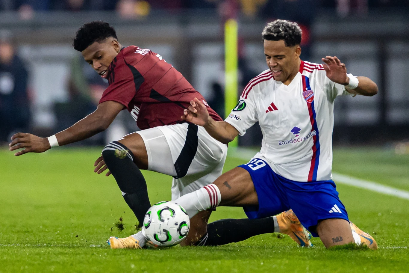 Michael Ameyaw (z prawej) kontra John Mercado Dwóch piłkarzy ściera się o piłkę na murawie podczas dynamicznej akcji meczowej, jeden ubrany w bordową koszulkę i czarne spodenki, drugi w białą koszulkę z niebieskimi spodenkami, widoczne jest skoncentrowanie i zaangażowanie zawodników.