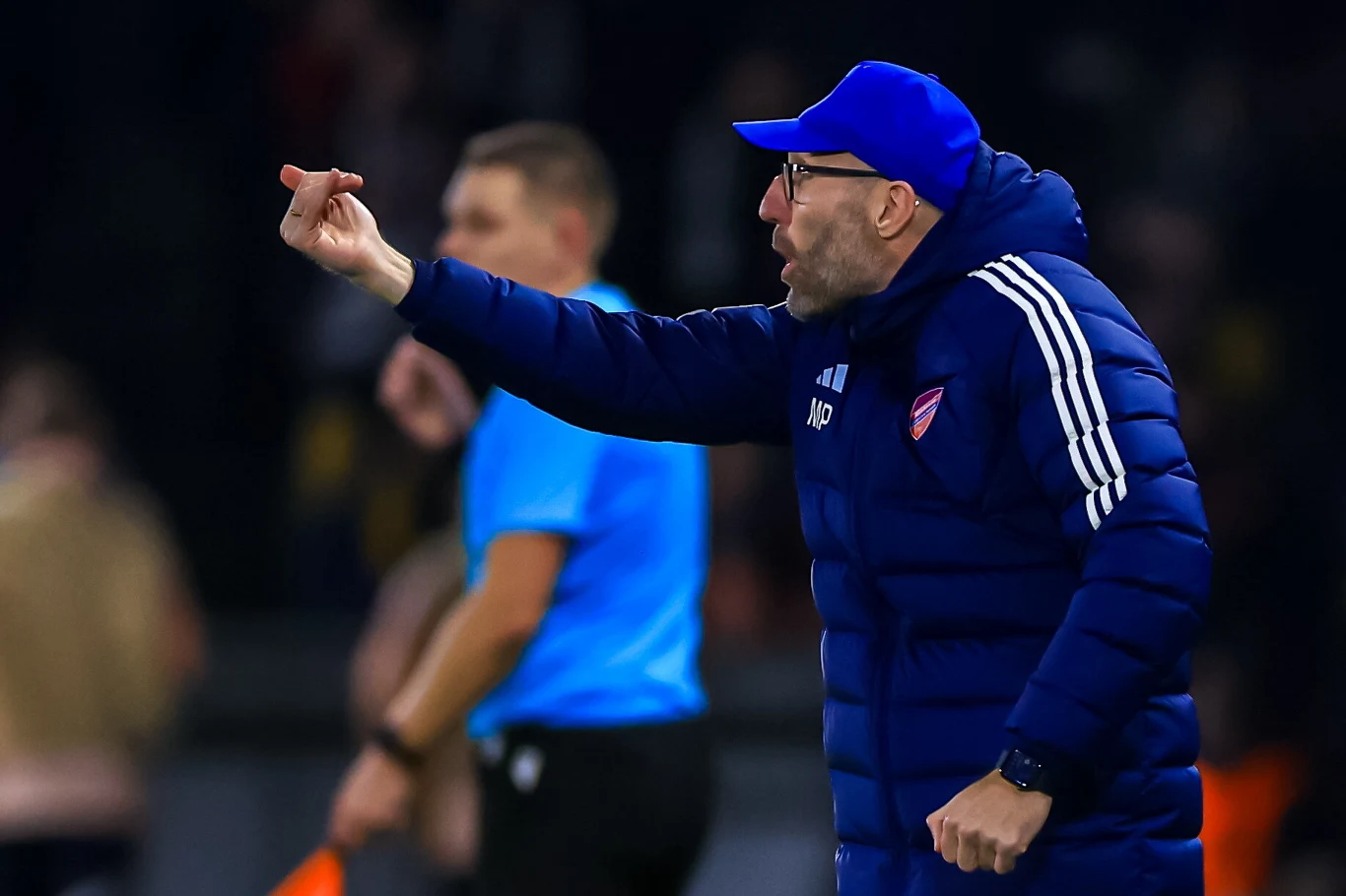 Trener Marek Papszun w roli dyrygenta Ubrany w sportową kurtkę i czapkę mężczyzna wykonuje gest środkowym palcem w kierunku boiska, w tle widoczny sędzia i rozmyte postacie.