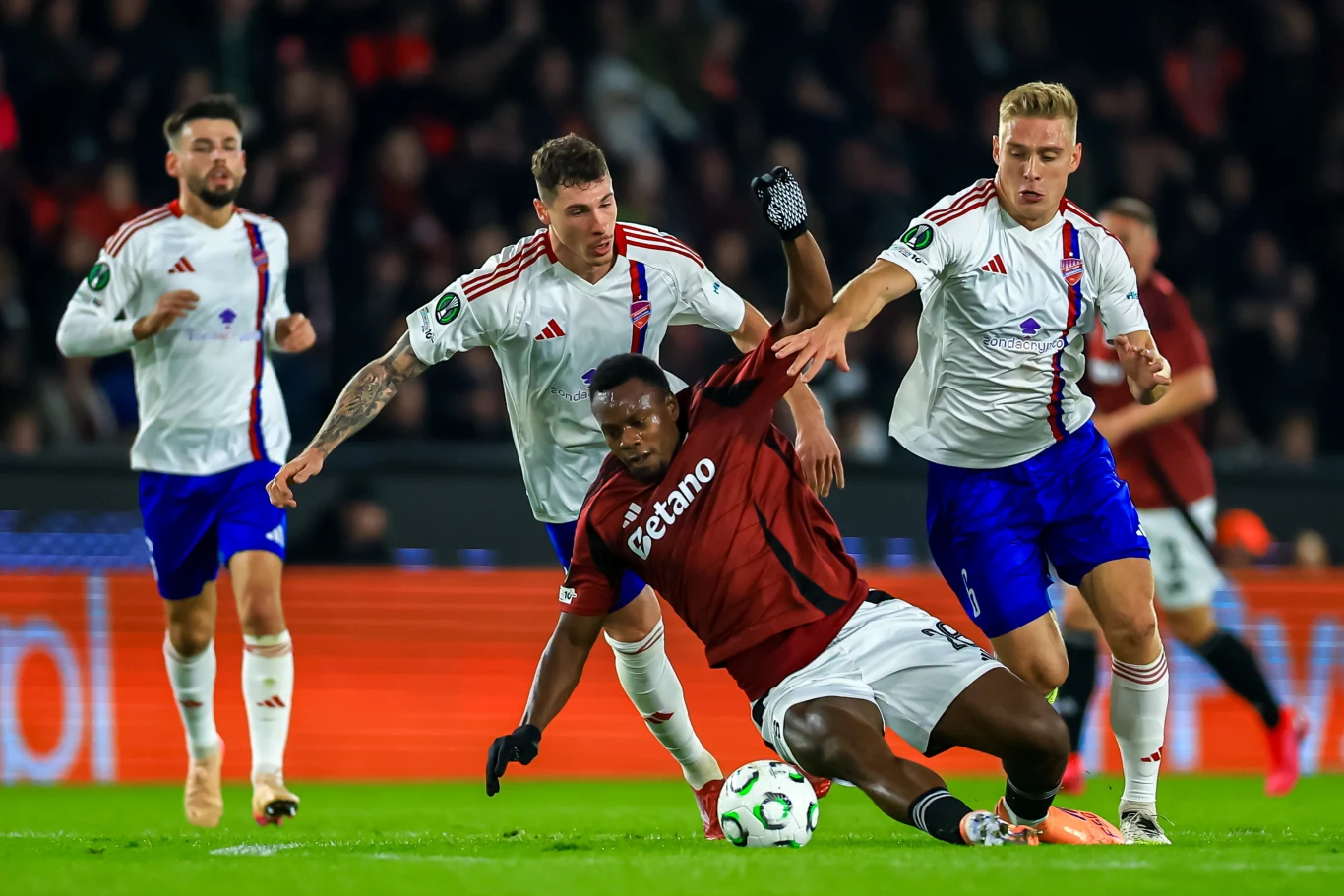 Kevin-Prince Milla w potrzasku Piłkarze dwóch drużyn rywalizują o piłkę na boisku podczas meczu piłkarskiego, jeden z zawodników upada na murawę, a trzech innych intensywnie walczy o przewagę.