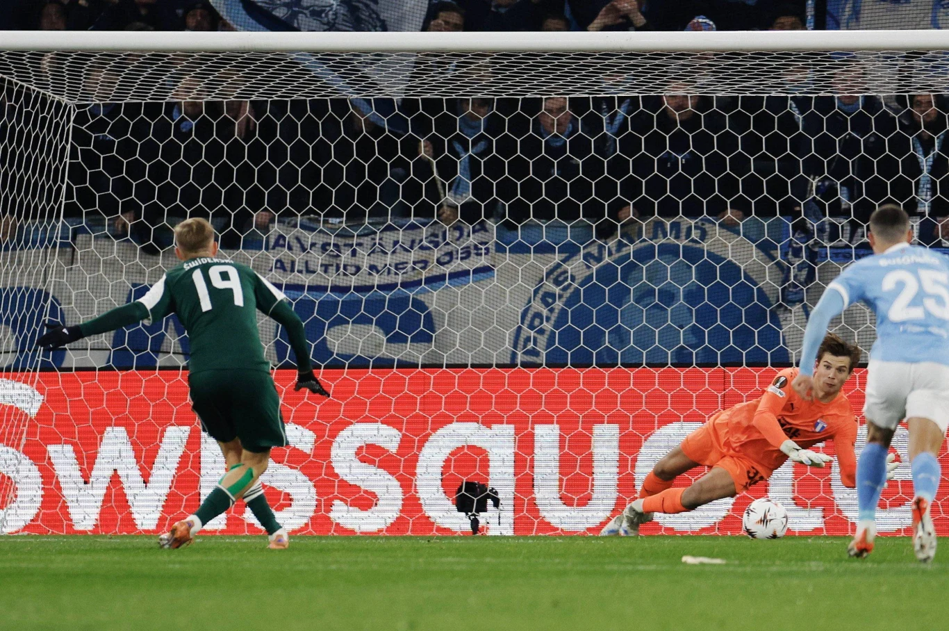 Karol Świderski nie wykorzystał rzutu karnego w meczu Ligi Europy ze szwedzkim Malmo Bramkarz ubrany w pomarańczowy strój rzuca się w kierunku piłki podczas obrony rzutu karnego, piłkarz w zielonej koszulce z numerem 19 biegnie po boisku, za nimi widoczny fragment bramki, siatki i kibice na trybunach.