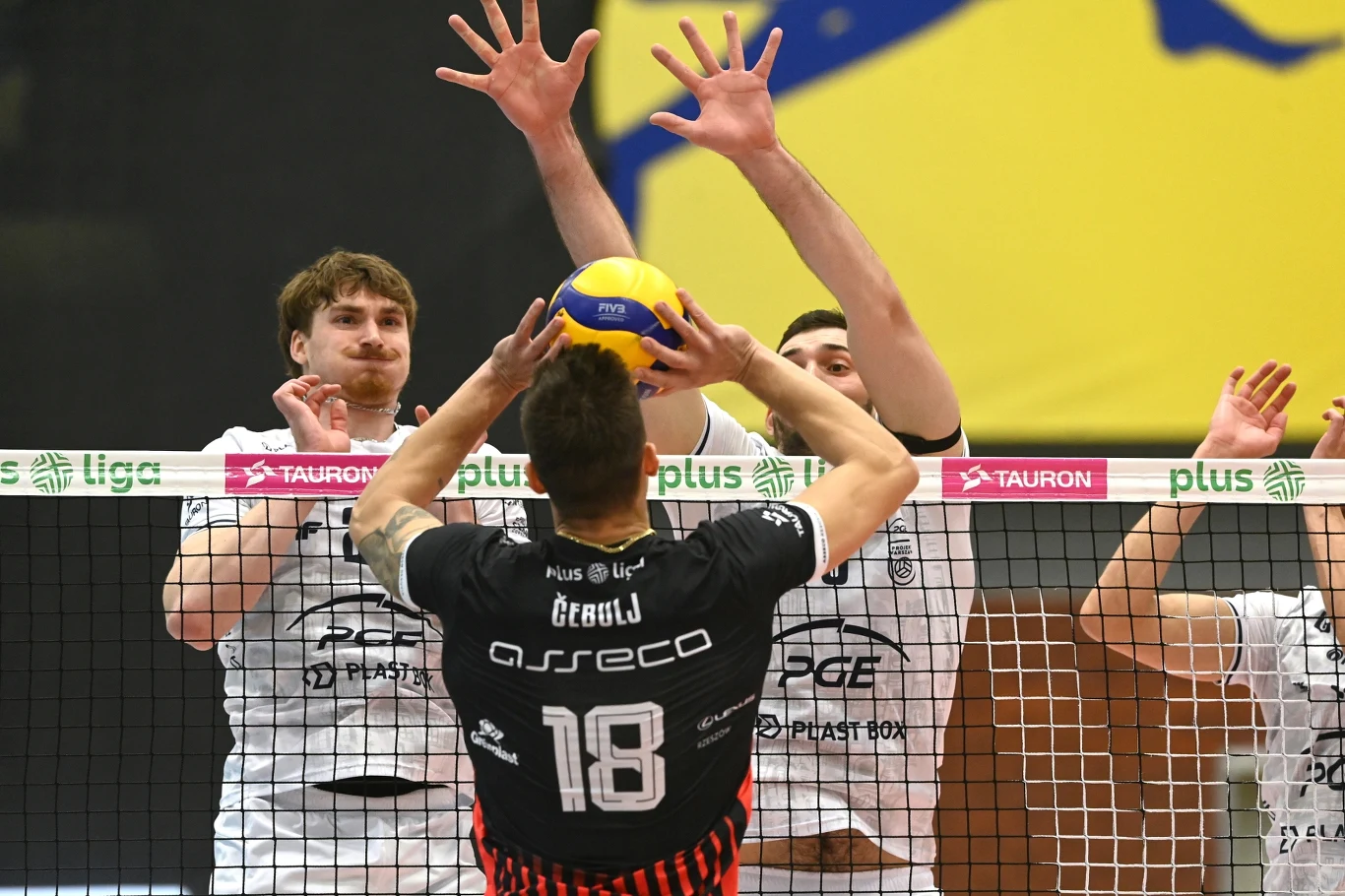 Dwóch siatkarzy w białych strojach próbuje zablokować atak zawodnika drużyny przeciwnej w czarnym stroju, który podbija piłkę przy siatce; w tle kontrastujący żółty baner.