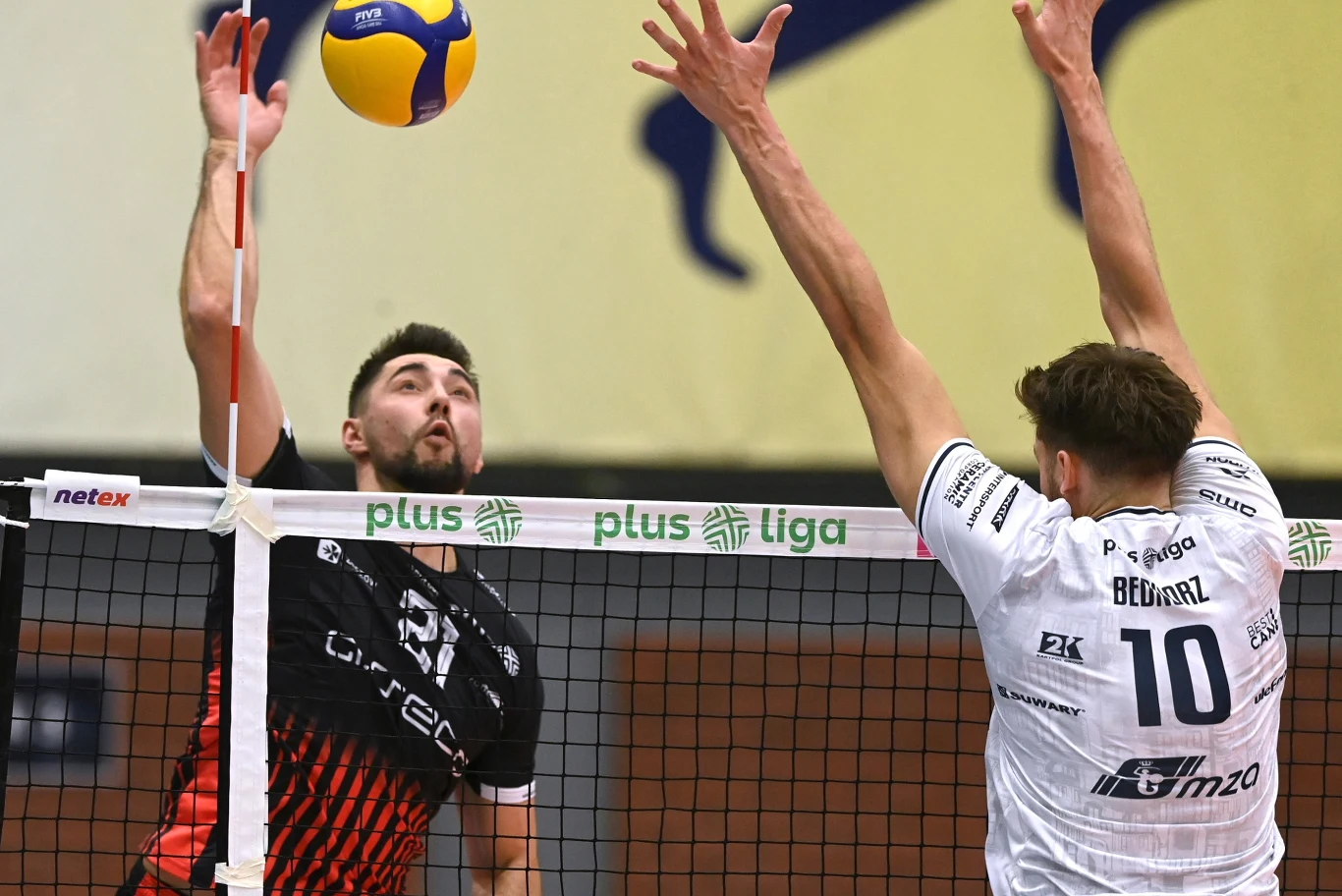 Dwóch siatkarzy rywalizuje przy siatce podczas meczu Plus Ligi. Zawodnik w czarnym stroju przygotowuje się do ataku piłki, a gracz w białej koszulce blokuje po drugiej stronie siatki. W tle widoczna jest siatka oraz fragmenty reklamy ligowej.