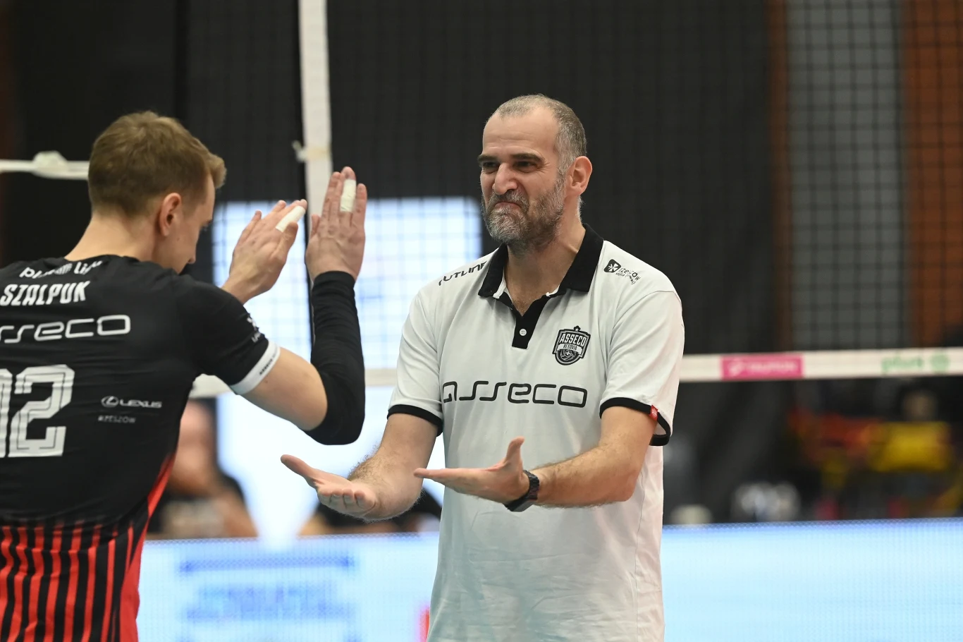 Trener w białej koszulce gestykuluje do zawodnika siatkówki w czarno-czerwonym stroju, obaj znajdują się po tej samej stronie siatki podczas meczu na hali sportowej.