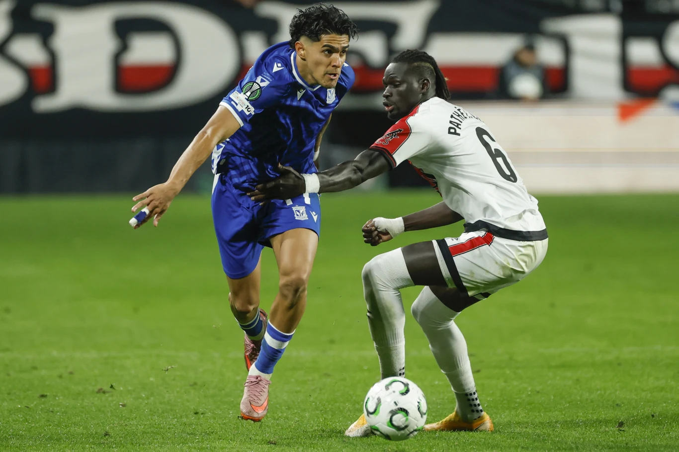 Luis Palma i Pathe Ciss Dwóch piłkarzy na murawie podczas walki o piłkę, jeden ubrany w niebieski strój sportowy, drugi w biało-czarny, intensywna akcja rozgrywająca się na tle trybun.