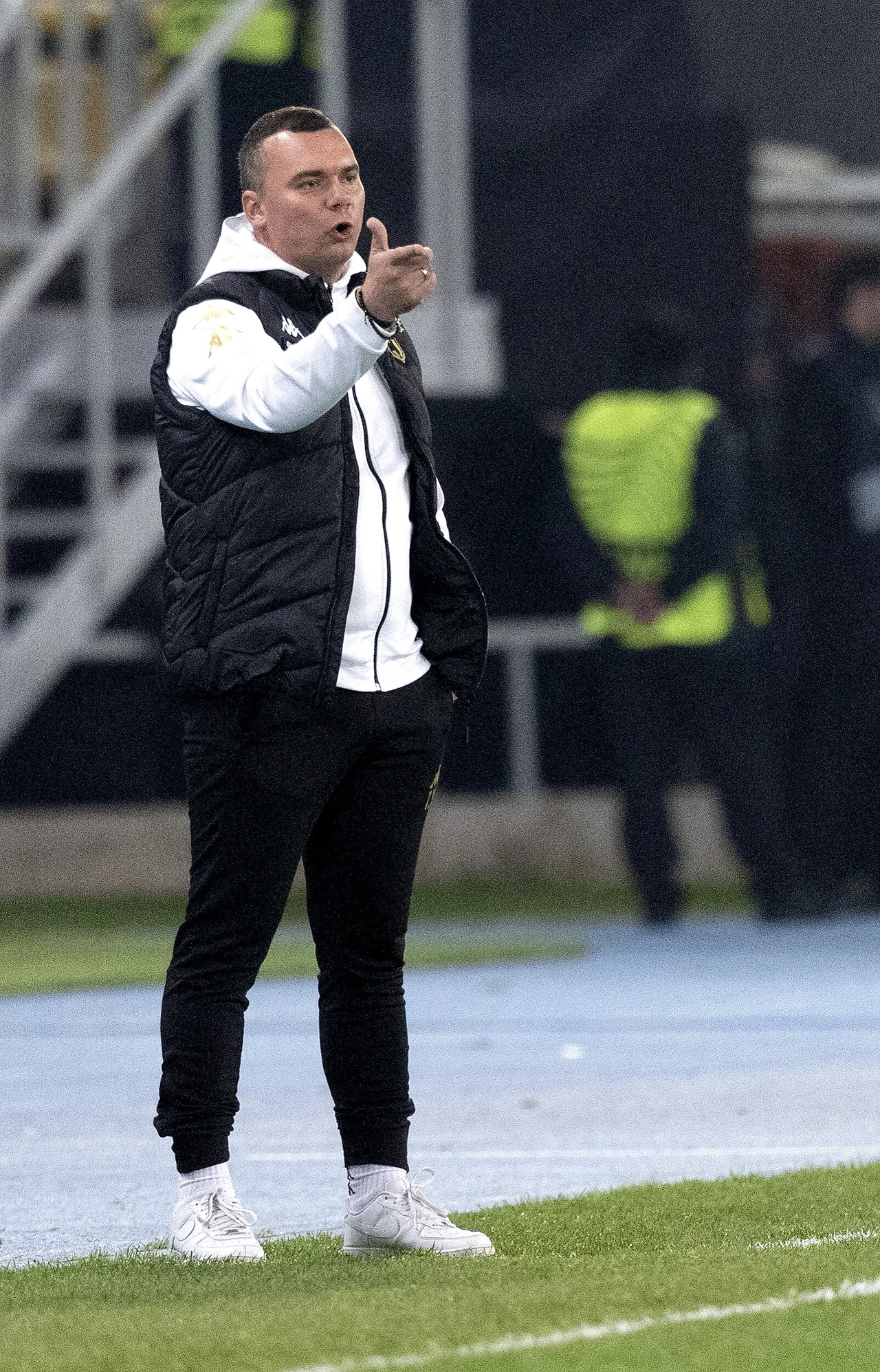 Mężczyzna w sportowej odzieży stoi przy linii bocznej boiska, wykonując gest dłonią i koncentrując wzrok przed siebie, w tle widoczni ochroniarze.