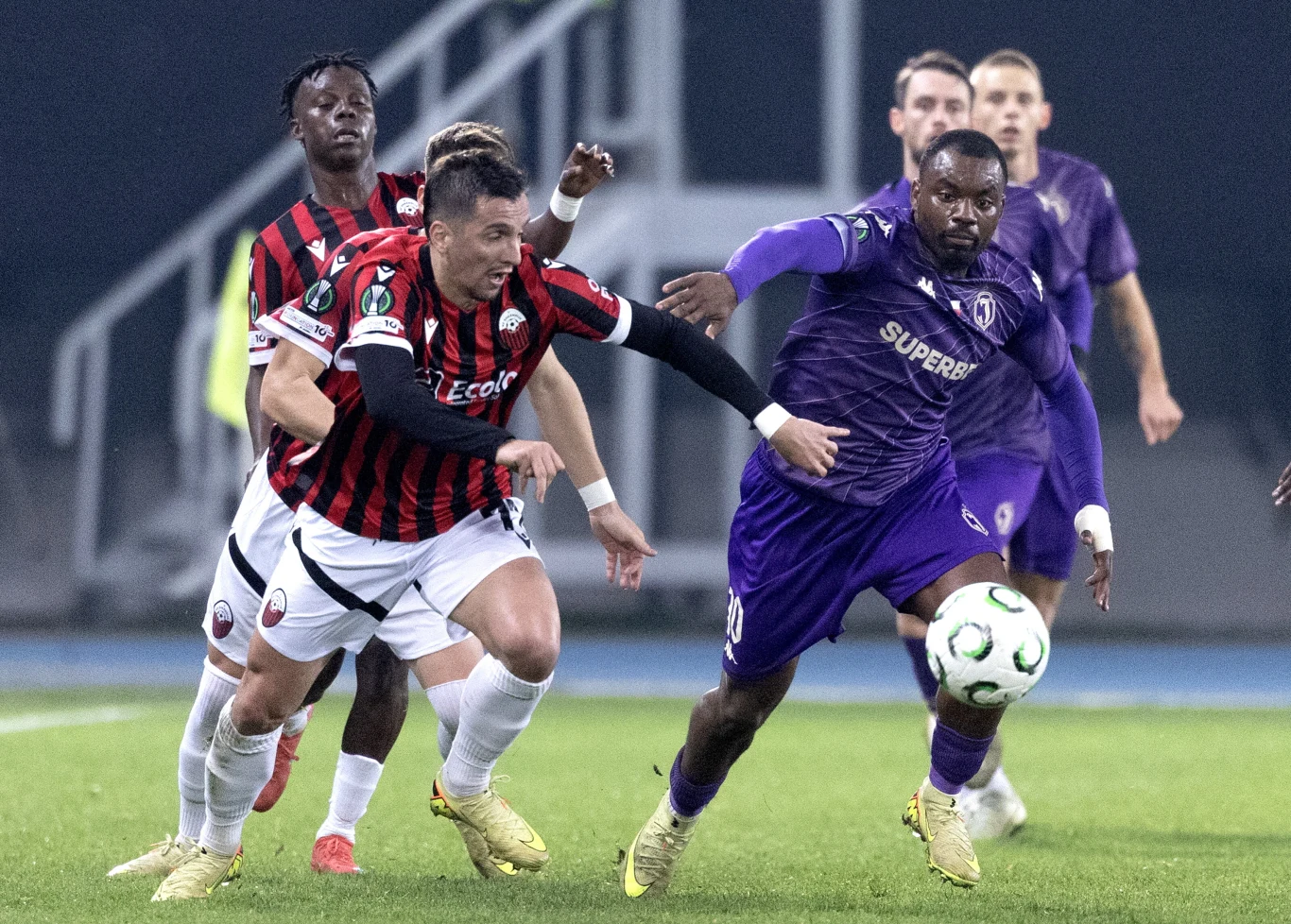 Piłkarze dwóch drużyn walczą o piłkę podczas dynamicznego meczu, jeden zawodnik w czarno-czerwonym stroju, drugi w fioletowym, pełne napięcia sportowe starcie na stadionie.