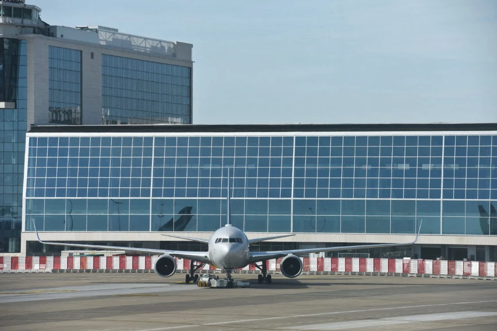Lotnisko w Brukseli Samolot pasażerski stoi na płycie lotniska przed nowoczesnym, przeszklonym terminalem. W tle widać dużą fasadę z oknami, a pas startowy jest wyznaczony barierkami i oznaczeniami.