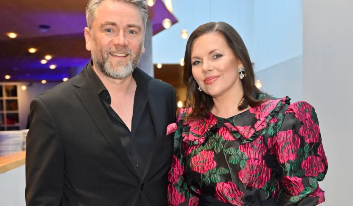 Kuba Badach i Aleksandra Kwaśniewska Elegancko ubrany mężczyzna w czarnym garniturze i kobieta w czarnej sukni w kolorowe kwiaty pozują razem, oboje uśmiechnięci, w tle jasne wnętrze i rozproszone światła.