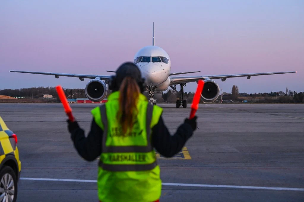 Pracownik obsługi naziemnej kieruje samolot na płycie lotniska za pomocą odblaskowych pałek świetlnych, naprzeciwko lądującej maszyny pasażerskiej. Widoczne są także elementy infrastruktury lotniska oraz samochód służbowy po lewej stronie.