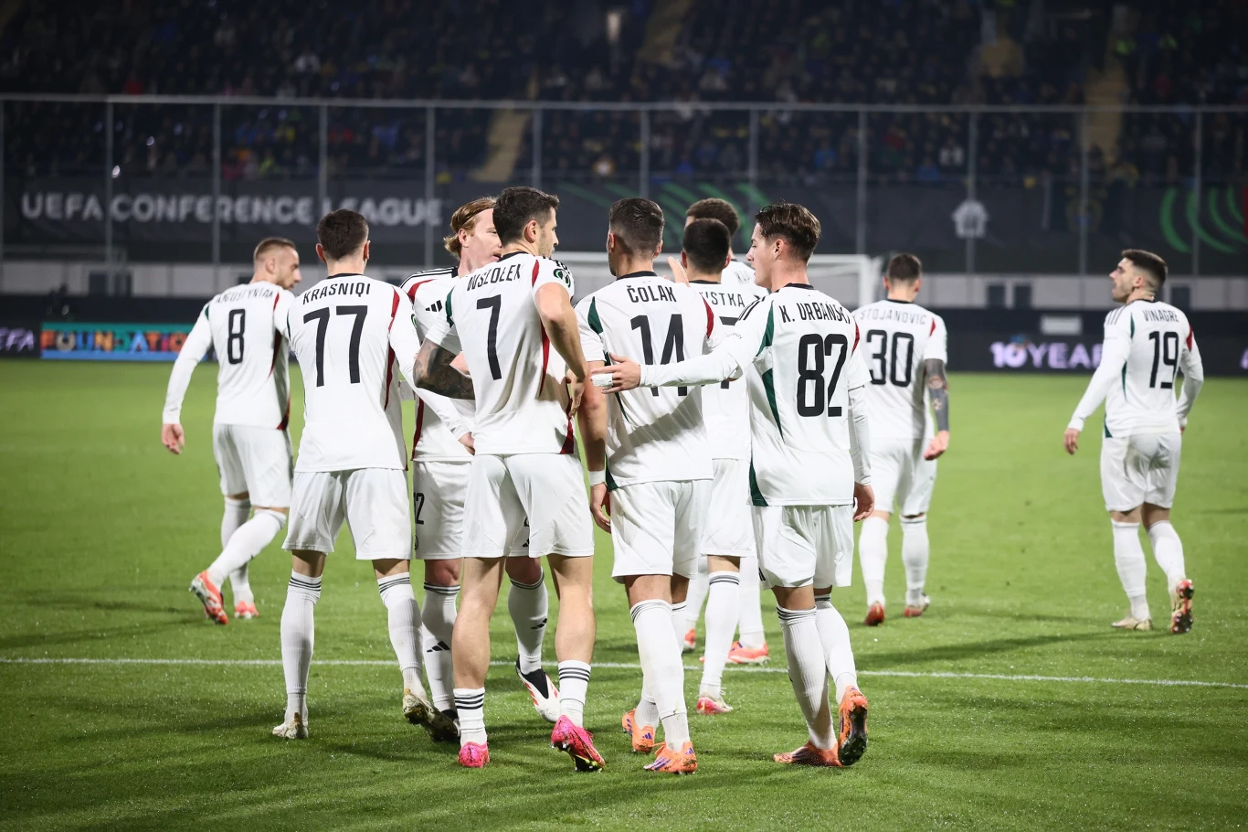 Piłkarze Legii Warszawa Grupa piłkarzy ubranych w białe stroje sportowe świętuje na boisku podczas meczu UEFA Conference League, wokół widoczne trybuny z kibicami oraz oświetlony stadion.