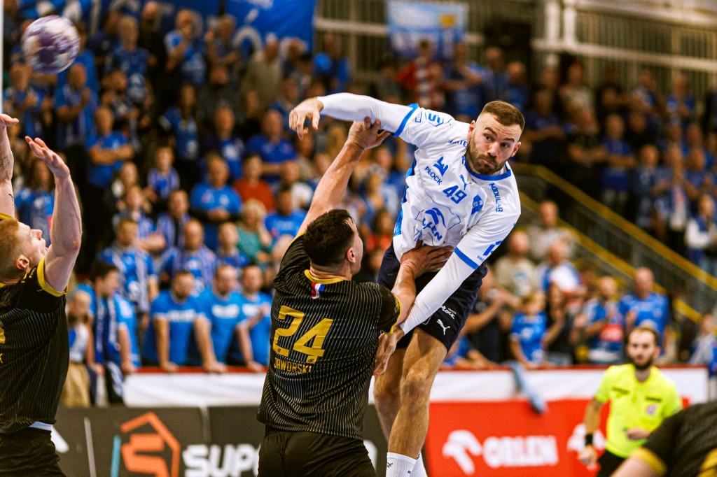 Piłkarz ręczny w białej koszulce skacze do rzutu w kierunku bramki, otoczony przez zawodników drużyny przeciwnej w czarnych strojach. W tle liczna widownia kibicująca na trybunach oraz sędzia obserwujący akcję.