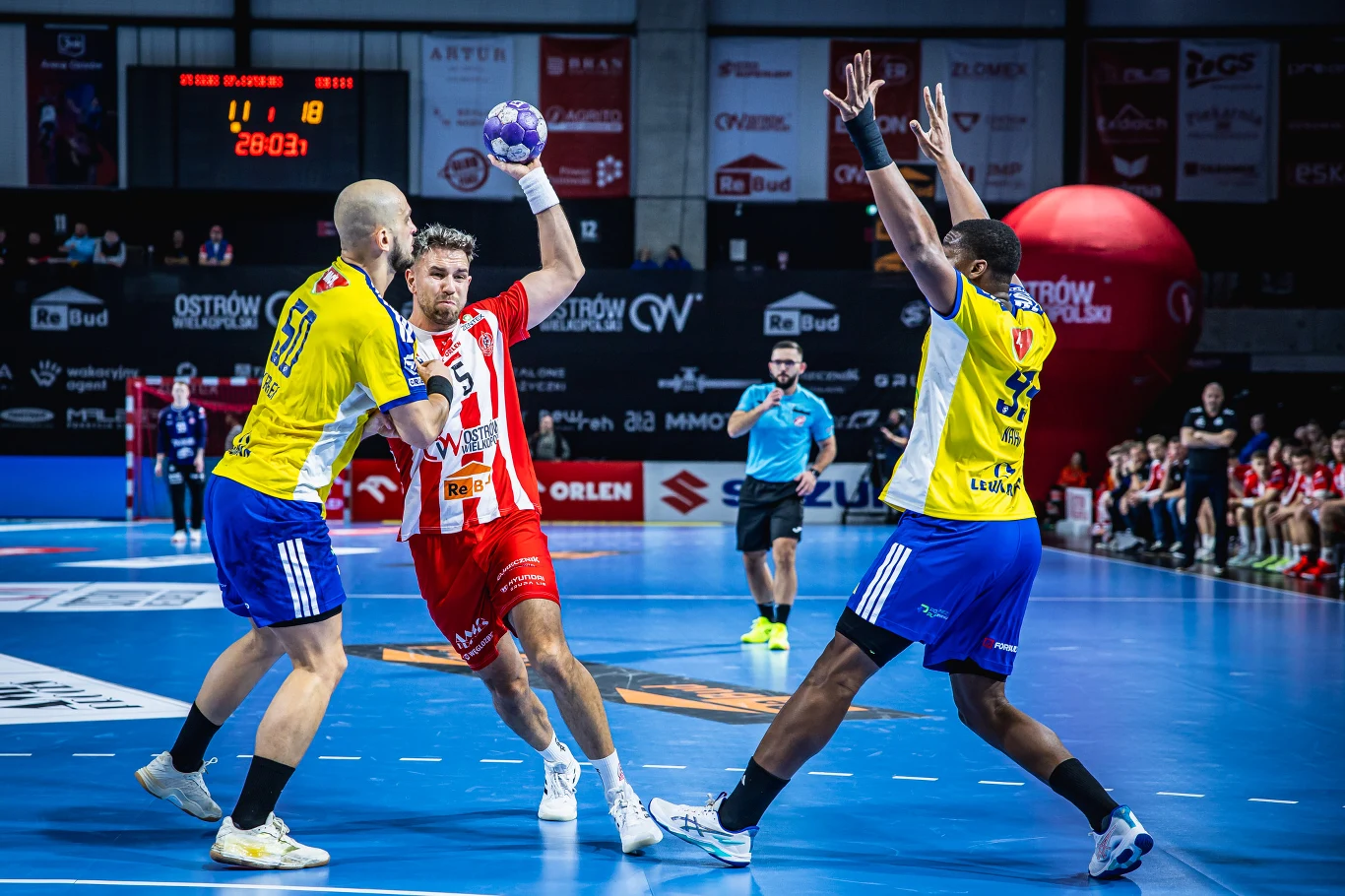 Zawodnik w czerwono-białym stroju stara się rzucić piłkę ręczną ponad dwoma obrońcami w żółto-niebieskich koszulkach na hali sportowej. Sędzia oraz pozostali zawodnicy i kibice obserwują akcję.