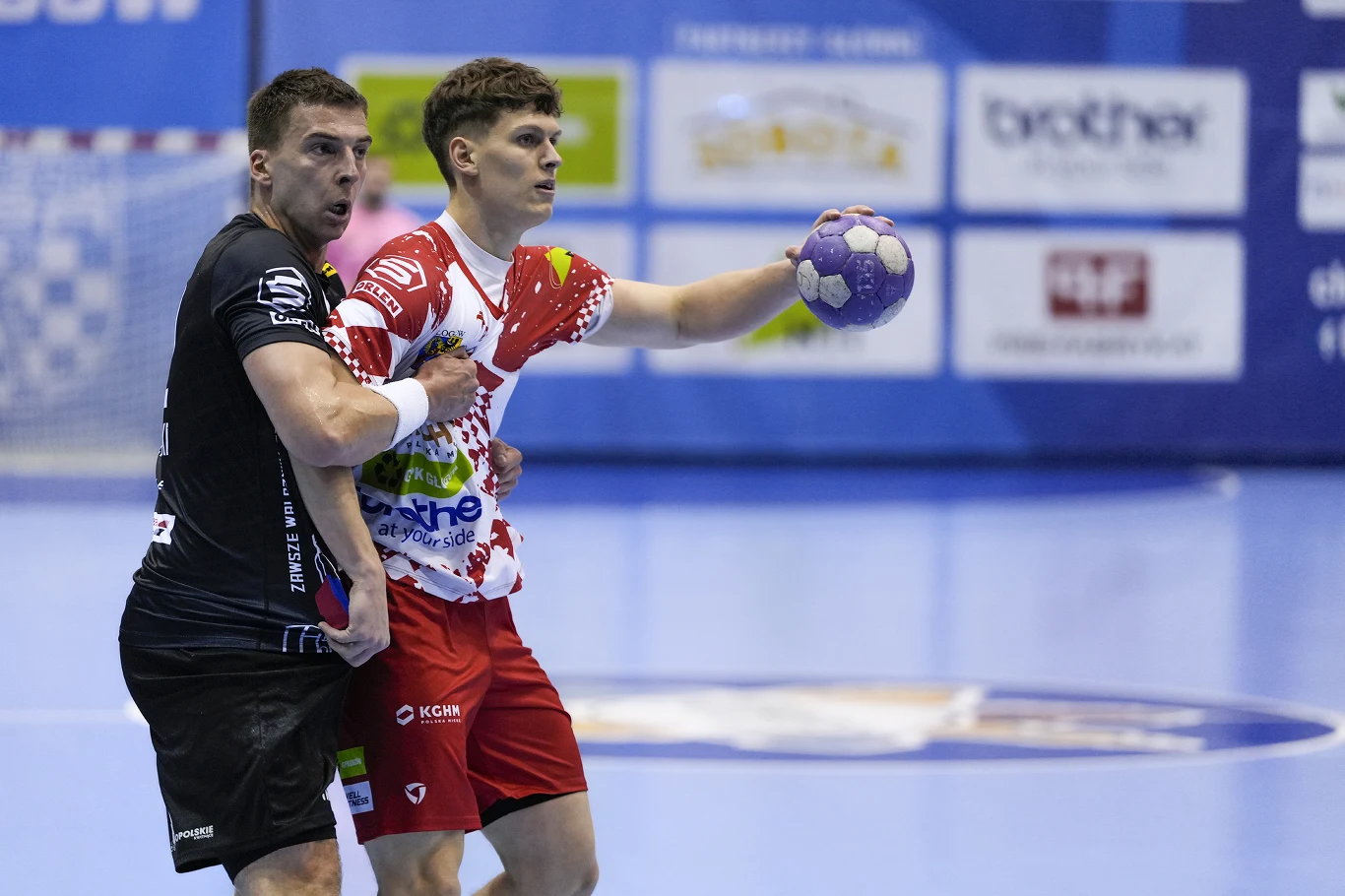 Dwóch zawodników podczas meczu piłki ręcznej, jeden w czarnej koszulce zatrzymuje drugiego, który trzyma piłkę w jednej ręce, tło stanowią reklamy oraz fragment boiska.