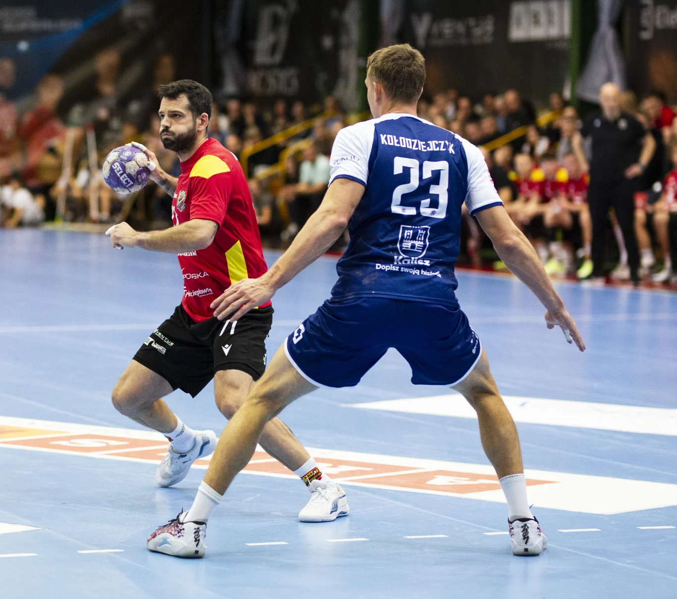 Dwóch zawodników podczas dynamicznej akcji meczu piłki ręcznej, jeden z nich w czerwonym stroju przygotowuje się do rzutu na bramkę, drugi w niebieskim stroju próbuje go zablokować, w tle widoczni kibice oraz ławka rezerwowych.