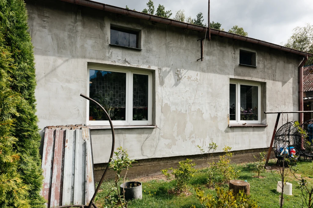 Szara, nieotynkowana elewacja domu jednorodzinnego z dwoma oknami i jednym małym oknem na górze, przed budynkiem ogród z roślinami, fragment starej blachy i metalowy stelaż po lewej stronie.