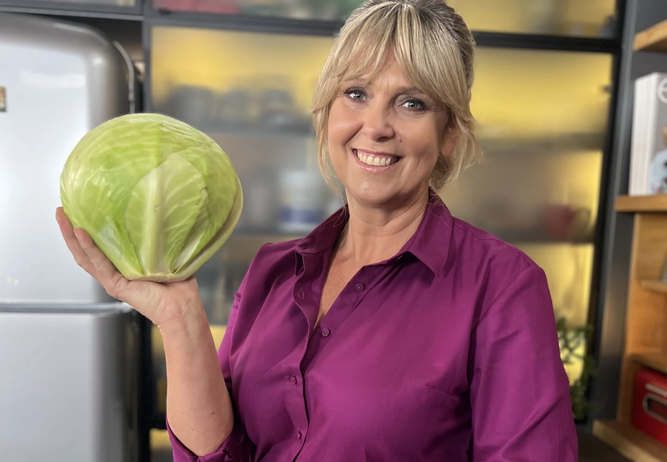 Ewa Wachowicz Uśmiechnięta kobieta w fioletowej koszuli trzyma w dłoni dużą główkę kapusty, stojąc w nowoczesnej kuchni z lodówką w tle.