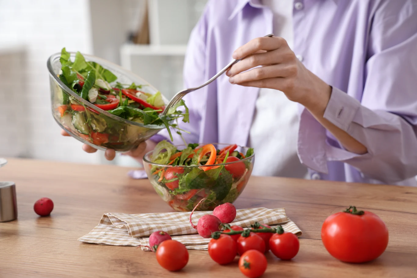 Osoba miesza świeżą sałatkę warzywną w szklanej misce, obok na stole leżą pomidory, rzodkiewki oraz ściereczka w kratkę.