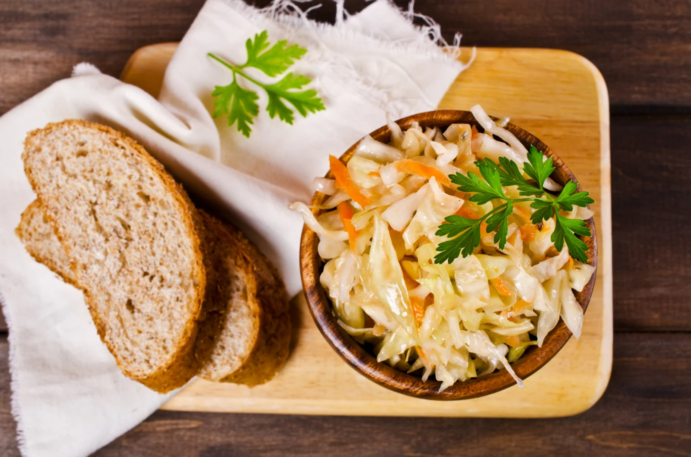 Coleslaw Drewniana deska z miską surówki z kapusty i marchewki, udekorowaną gałązką natki pietruszki, obok leżą trzy kromki chleba na białej serwetce.