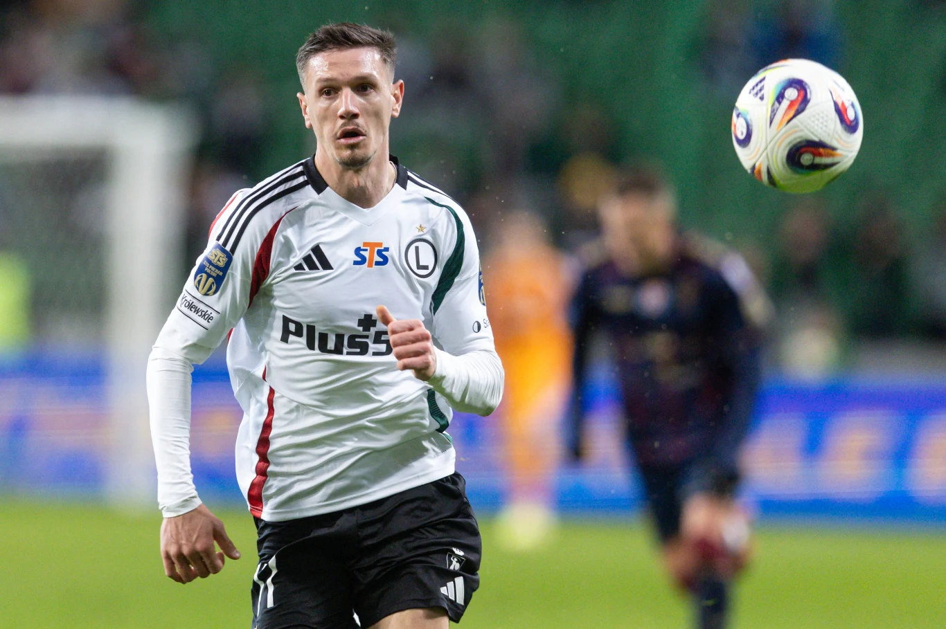 Ermal Krasniqi z Legii Warszawa Piłkarz w biało-czarnej koszulce Legii Warszawa biegnie za lecącą piłką podczas meczu piłkarskiego, w tle widoczni są inni zawodnicy na boisku oraz rozmyte trybuny.