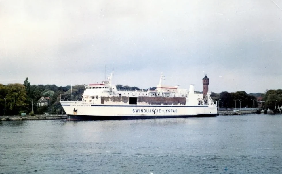 Prom pasażerski płynący po wodzie, z napisem Świnoujście – Ystad na burcie, w tle zieleń drzew oraz ceglana wieża latarni morskiej.