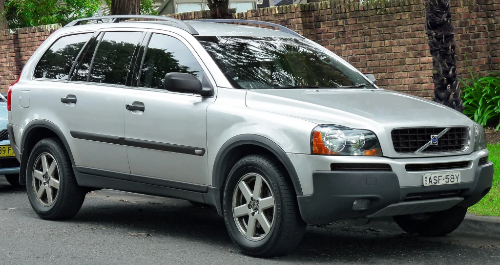 Srebrny SUV marki Volvo XC90 zaparkowany przy krawężniku na tle ceglanego muru i zieleni.