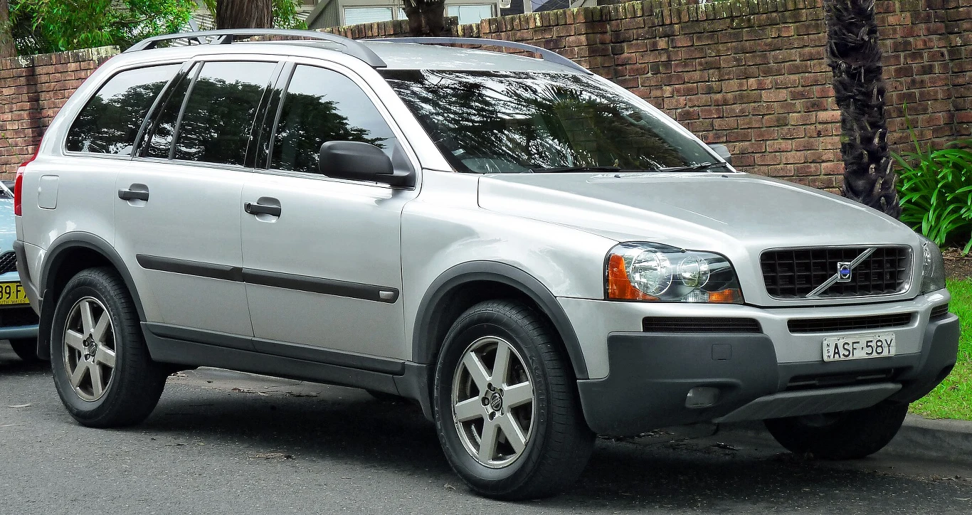 Srebrny SUV marki Volvo XC90 zaparkowany przy krawężniku na tle ceglanego muru i zieleni.