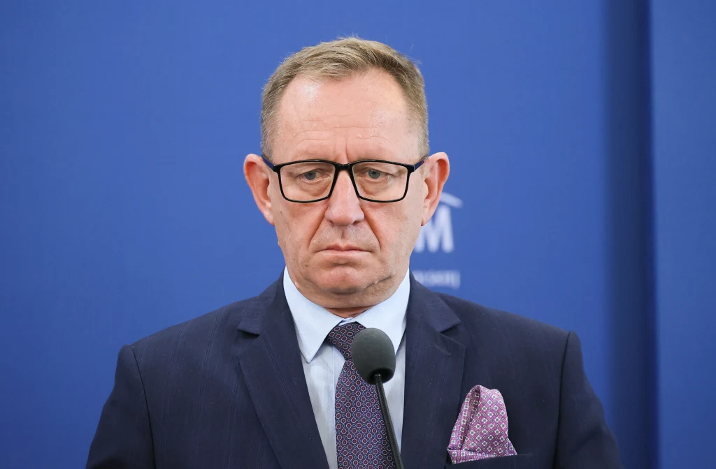 Bolesław Piecha wzywa Roberta Telusa do przeproszenia za aferę z działką pod CPK Mężczyzna w okularach w garniturze i krawacie stoi przed niebieskim tłem z mikrofonem, zachowując poważny wyraz twarzy.